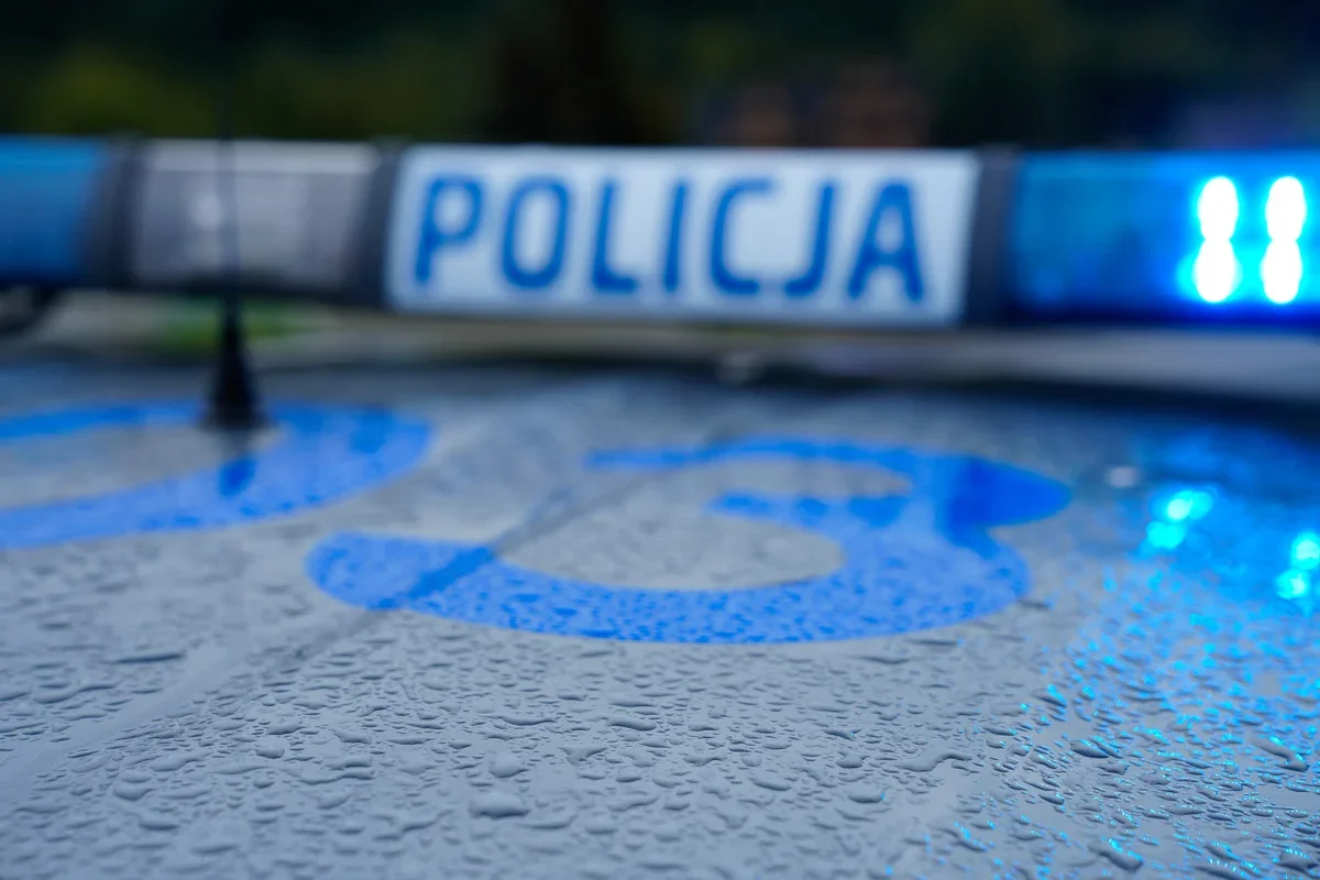 Zbliżenie na dach policyjnego radiowozu z napisem „POLICJA” i błękitnym światłem, pokryty kroplami deszczu.