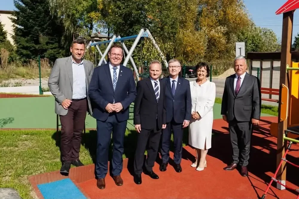 Grupa sześciu osób w eleganckim stroju stoi na kolorowej nawierzchni placu zabaw z huśtawkami i zielenią w tle.