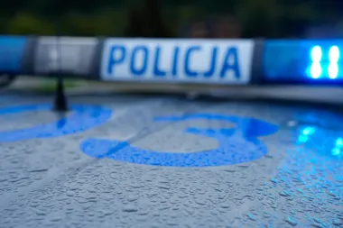 Zbliżenie na dach policyjnego radiowozu z napisem „POLICJA” i błękitnym światłem, pokryty kroplami deszczu.