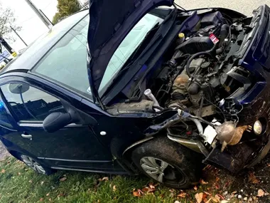 Uszkodzony ciemny samochód osobowy stoi na poboczu trawiastym przy drodze, z otwartą maską i drzwiami, po zderzeniu czołowym.