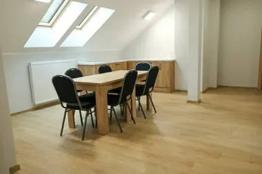 Pusty, jasny pokój na poddaszu z drewnianym stołem i czarnymi krzesłami, pod oknami dachowymi, z drewnianą zabudową w tle.