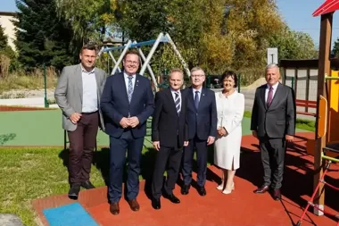 Grupa sześciu osób w eleganckim stroju stoi na kolorowej nawierzchni placu zabaw z huśtawkami i zielenią w tle.