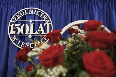Dekoracja z okazji 150-lecia szkolnictwa w Leńcach, z czerwonymi różami na tle niebieskiej kotary z okrągłym emblematem.