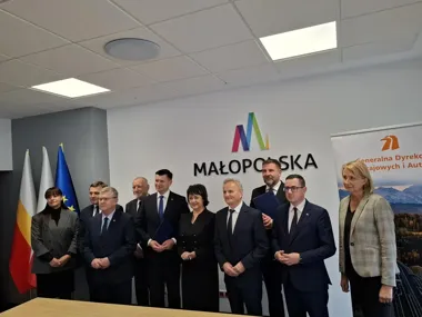 Grupa osób w formalnym stroju stoi podczas uroczystości w siedzibie urzędu województwa małopolskiego, w tle logo Małopolski i flagi.